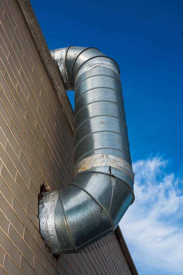 services-02 A large industrial air duct attached to a brick wall under a bright blue sky.